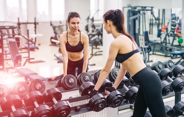 Mooi meisje met handen op halters kijken op haar reflectie in de spiegel in een sporthal slanke jonge vrouw neemt halters voor een spiegel in fitnessclub