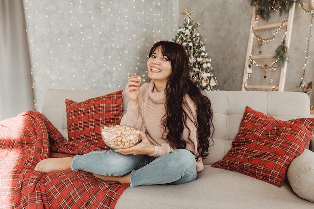 Mooi meisje met lang donker haar ligt op de bank en eet popcorn op de achtergrond van de kerstboom.