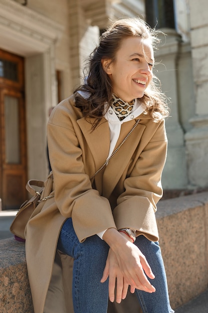 Mooi meisje met lang haar in beige jas op een zonnige dag zittend op een stoeprand en glimlachen. Streetstyle portret