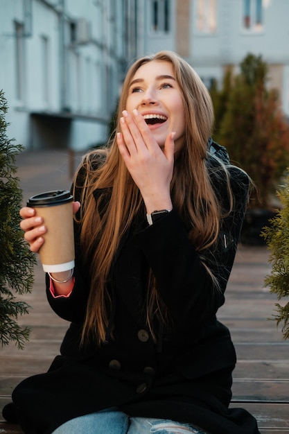 Mooi meisje met lang haar met koffie zittend op een bankje en lachen Mooie dame in een jas streetstyle portret