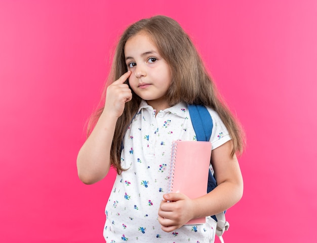 Mooi meisje met lang haar met rugzak met notitieboekje wijzend met wijsvinger naar haar oog kijkend naar de voorkant met serieus gezicht over roze muur