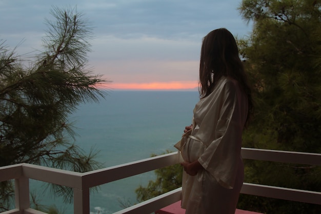 mooi meisje met lang haar, staande in een witte jas op het balkon van het hotel met uitzicht op de zee en de zonsondergang