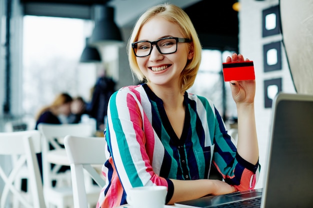 Foto mooi meisje met lichte haren dragen kleurrijke shirt en bril zitten in café met laptop en creditcard, freelance concept, online winkelen, glimlachend.