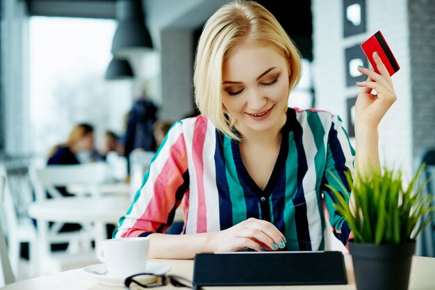 Mooi meisje met lichte haren dragen kleurrijke shirt zitten in café met tablet, creditcard en kopje koffie, freelance concept, online winkelen.