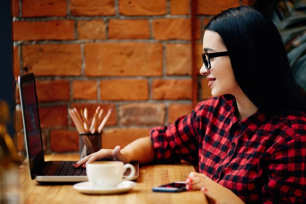 Mooi meisje met zwart haar dragen bril zitten in café met laptop en kopje koffie, freelance concept, portret, rode shirt dragen.