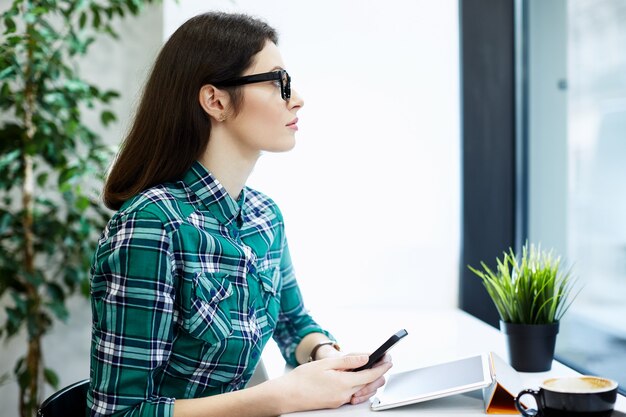 Mooi meisje met zwart haar dragen shirt en bril zitten in café met tablet en kopje koffie, freelance concept, venster kijken.