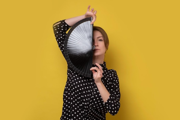 Mooi meisje met zwarte ventilator die de helft van haar gezicht verbergt Studio-opname op gele muur
