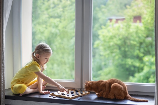 Mooi meisje op de vensterbank speelt dammen met een kat