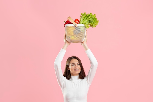 Mooi meisje rekt een mand met glasvezel van gezonde groenten uit op een roze achtergrond
