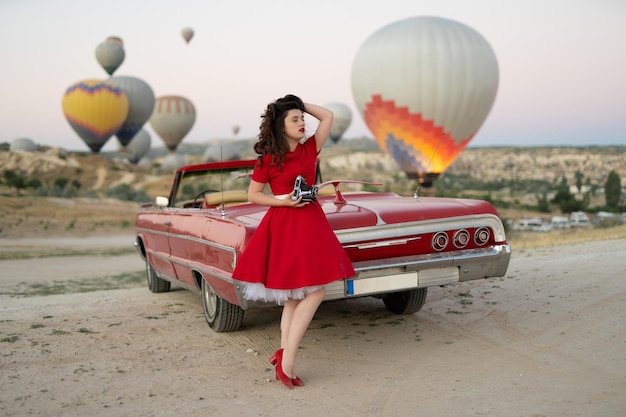 Mooi meisje retro-stijl poseren in de buurt van vintage rode cabriolet auto met oude filmcamera op de achtergrond van ballonnen in Cappadocië