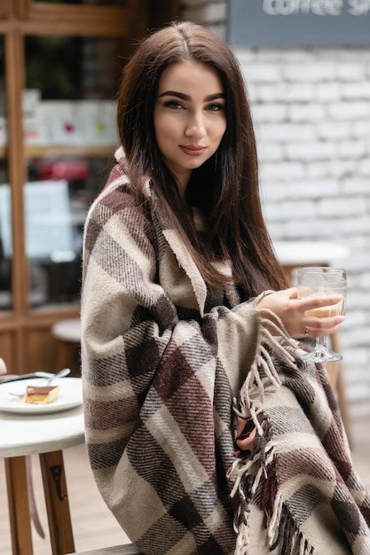 Mooi meisje rusten en koffie drinken zittend in café op het terras, gewikkeld in een wollen geruite deken. Herfst ochtend. Mooie jonge vrouw met kopje koffie buiten op herfstdag