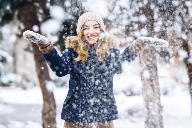 Mooi meisje speelt met sneeuw in het winterbos Glimlachend meisje in een blauwe jas en gebreide muts