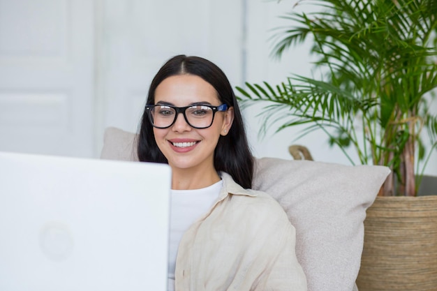 Mooi meisje studeert online thuis zittend op de bank in de woonkamer brunette in glazen