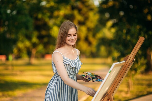 Mooi meisje tekent een foto in het park met behulp van een palet met verf en een spatel. Schildersezel en canvas met een afbeelding.