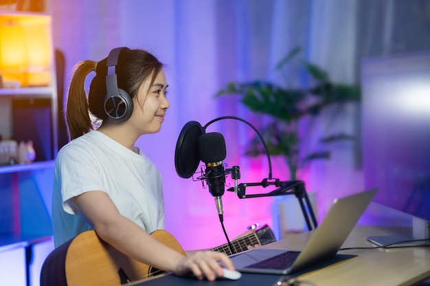 Mooi meisje zingt met hoofdtelefoon en speelt gitaar en neemt nieuw nummer op met microfoon in de thuisopnamestudio