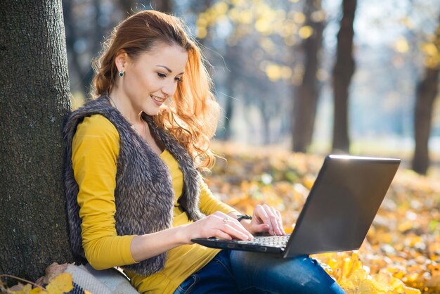 Mooi meisje zit onder een boom tussen de gele bladeren en gebruikt een laptop in het herfstpark