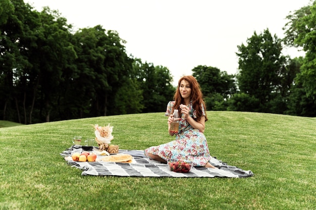 Mooi meisje zit op een deken in het park en houdt een drankje in haar handen. meisje met sproeten en rood haar op een picknick. ecologisch voedsel. zelfgemaakte producten.