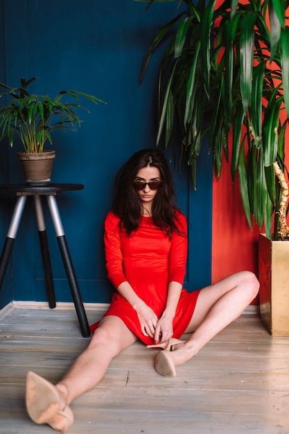 Mooi mode slank meisje met donker lang haar, in rode elegante jurk en zonnebril die zich voordeed op blauwe rode muur in de studio. Indor soft focus portret van stijlvolle brunette babe zit tussen twee planten.