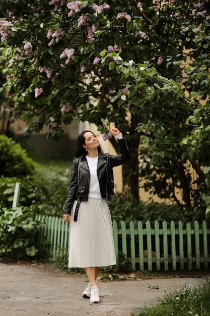 Mooi model meisje in een witte midi rok en zwarte leren jas poseren in de buurt van een bloeiende lila boom jonge brunette vrouw in lente outfit poseren buitenshuis