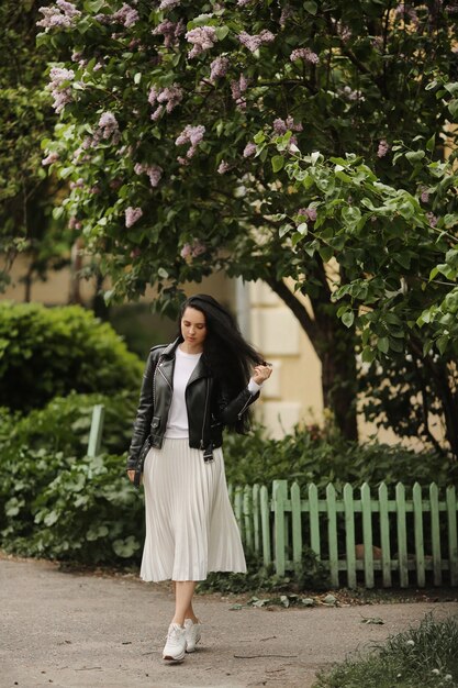 Mooi model meisje in zomer midi rok en leren jas poseren in de buurt van een bloeiende boom jonge brunette vrouw in lente outfit poseren buitenshuis