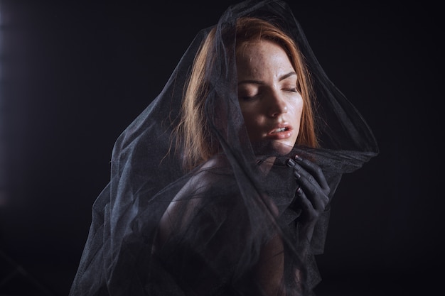 Mooi model met rood haar poseren achter een zwarte stof en een sluier in een studio