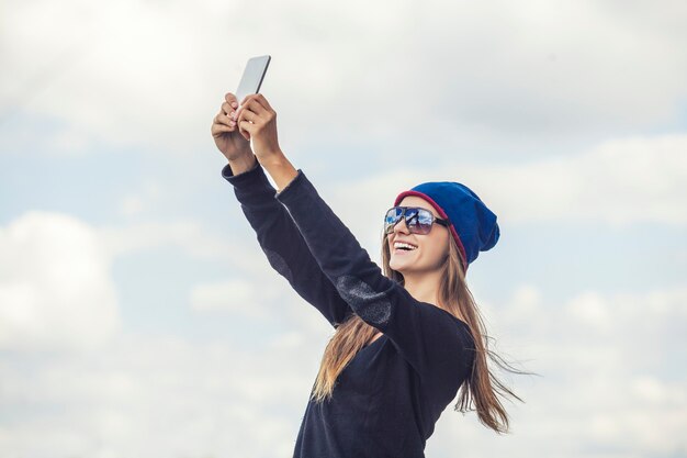 Mooi model vrouw met de telefoon tegen de hemel maakt selfie. Communicatie, fotografie, portret, sociaal, stijl