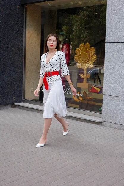 Mooi model ziet er brunette vrouw uit in een witte jurk met zwarte stippen terwijl ze wakker wordt op de achtergrond van een stadsstraat met stijlvolle tassen in de hand.