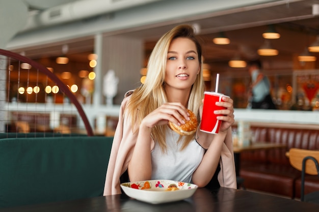 Mooi modieus gelukkig meisje met een hamburger en cola fastfood eten in een café