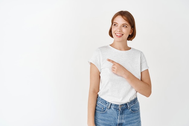 Mooi natuurlijk meisje zonder make-up witte tanden glimlachend en wijzend naar links naar het logo met banner die in een t-shirt op de achtergrond van de studio staat