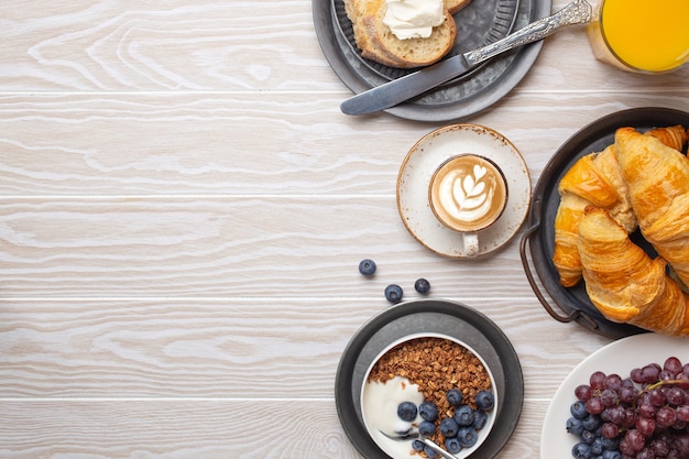 Mooi ochtendontbijt met verse croissants, muesli met yoghurt en bessen, toast, koffiecappuccino en fruit op plaat. lekker en gezond ontbijt op witte houten tafel, ruimte voor tekst