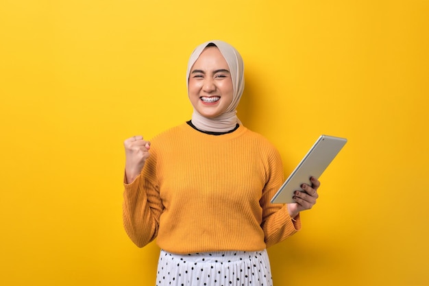 Mooi opgewonden Aziatisch meisje dat hijab draagt met behulp van digitale tablet die vuist opheft om succes te vieren dat op gele achtergrond is geïsoleerd