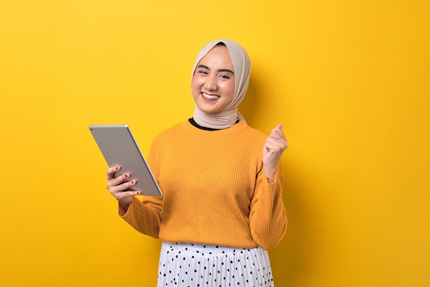 Mooi opgewonden Aziatisch meisje dat hijab draagt met behulp van digitale tablet die vuist opheft om succes te vieren dat op gele achtergrond is geïsoleerd