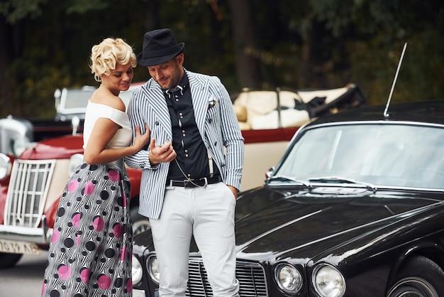 Foto mooi paar in ouderwetse slijtage in de buurt van retro auto met bos op de achtergrond.