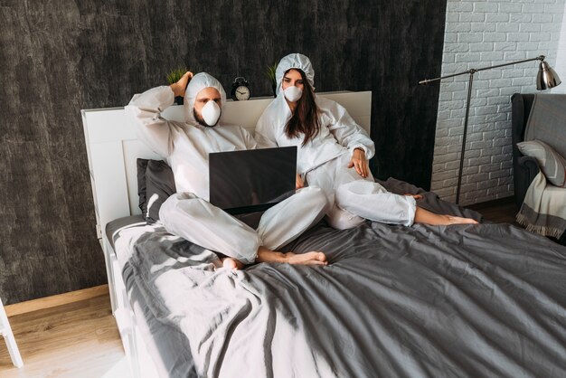 Mooi paar in witte beschermende kostuums met kappen en gezichtsmaskers thuis op het bed