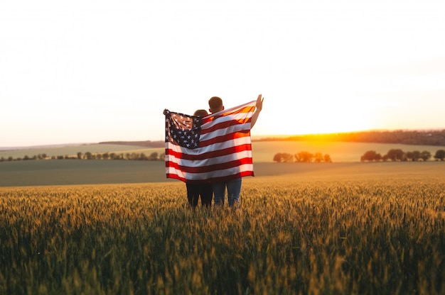 Foto mooi paar met de amerikaanse vlag in een tarweveld bij zonsondergang. independence day, 4 juli.