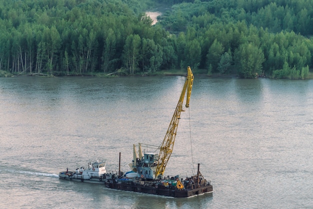 Mooi rivierlandschap met groene kusten en schip met exemplaarruimte. de sleepboot sleept aak met kraan langs rivieroever. dawn gereflecteerd op kalm wateroppervlak.