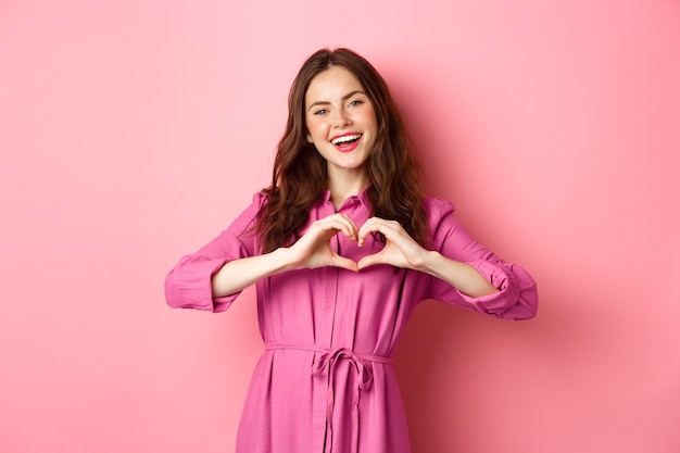 Mooi romantisch meisje glimlachen, hartgebaar tonen, staande in mooie jurk tegen roze muur.