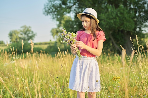 Mooi romantisch meisje in hoed, jurk verzamelen boeket van wilde bloemen in zonnige weide, pittoresk landschap, gouden uur, kopieer ruimte. Jeugd, zomer, natuur, schoonheid, kinderconcept.