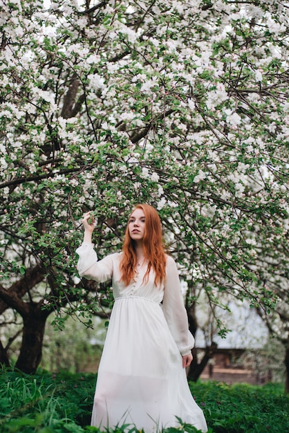 Mooi roodharig meisje in een witte jurk onder bloeiende appelbomen in de tuin