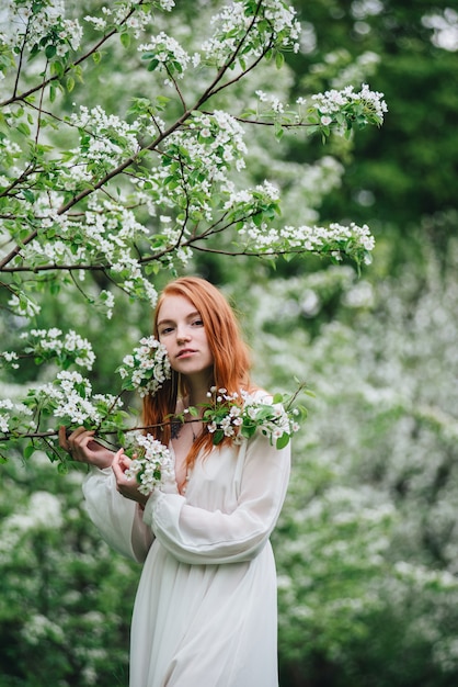 Mooi roodharig meisje in een witte jurk onder bloeiende appelbomen in de tuin