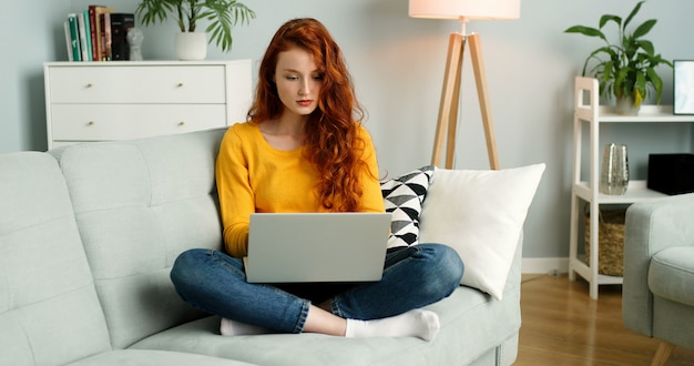 Mooi roodharigemeisje die zilveren laptop met behulp van terwijl thuis het zitten op bank in woonkamer