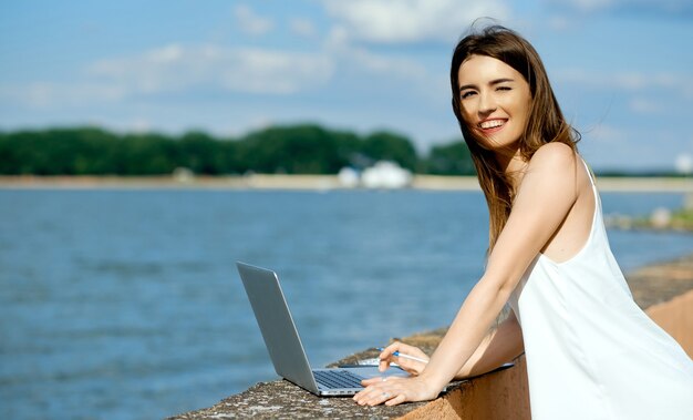 Mooi, serieus meisje in een witte jurk met een tablet, pen, laptoptelefoon op het dok. Hoge kwaliteit foto