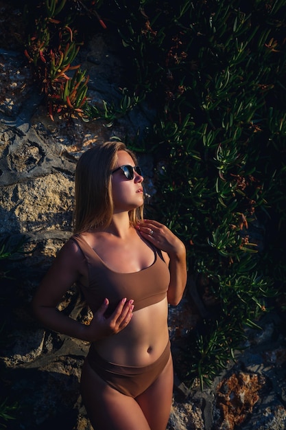 Mooi sexy meisje in zonnebril in een beige zwembroek Jonge vrouw met gebruind lichaam in de buurt van stenen muur met groene planten