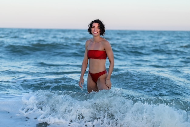 Mooi sexy meisje op het strand van de zee in een rode jurk kijkt naar de camera haren wapperen wind bij zonsondergang. Close-up van een modieus naakt meisje in een zwembroek. Close-up mode portret van model buiten