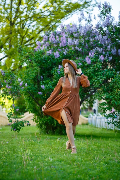 Mooi slank langharig meisje in een hoed op een achtergrond van lila bloemen. Ze is van Europese uitstraling met een glimlach op haar gezicht. Jonge vrouw loopt op een warme zonnige dag in een bloemrijk park