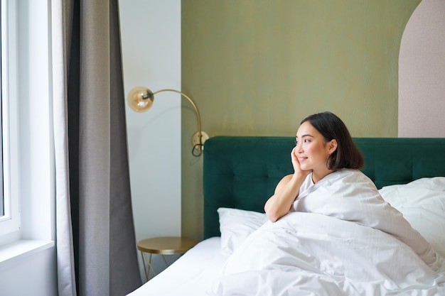 Mooi slaperig aziatisch meisje wordt wakker in haar bed met knusse witte dekens die naar buiten kijken met een tevreden glimlach Lifestyle concept