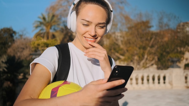 Mooi sportief meisje in koptelefoon met rugzak en voetbal gelukkig met smartphone onderweg