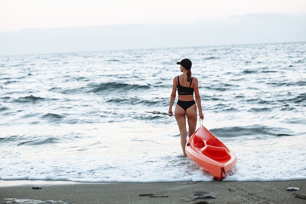 Mooi sportmeisje in een zwart zwempak gaat met een oranje kajak om in de zee te zwemmen bij zonsondergang