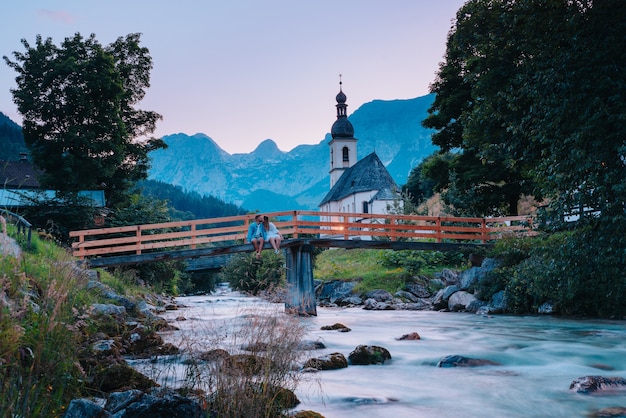 Mooi stel zittend op een brug over een rivier met bergen en een kerk op de achtergrond, in Ramsau, Duitsland in Berchtesgaden.