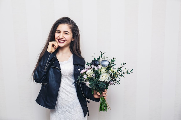 Mooi stijlvol meisje gekleed in leren jas, boeket bloemen te houden en poseren tegen witte muur.